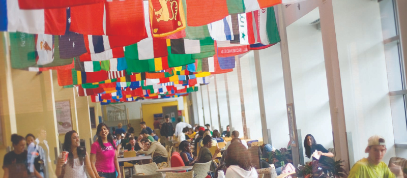 A large room of college students eat together beneath a ceiling full of international flags.