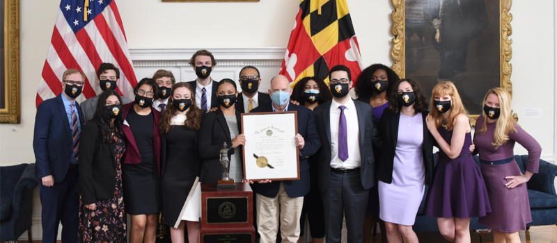A group of student men and women in business casual clothing pose with Maryland Governor Larry Hogan to celebrate their Mock Trial win. They are holding up a large certificate at the center of the group.