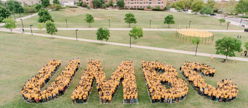 Hundreds of students in gold-colored t-shirts gather on a field to create the shape 'UMBC' with their bodies.